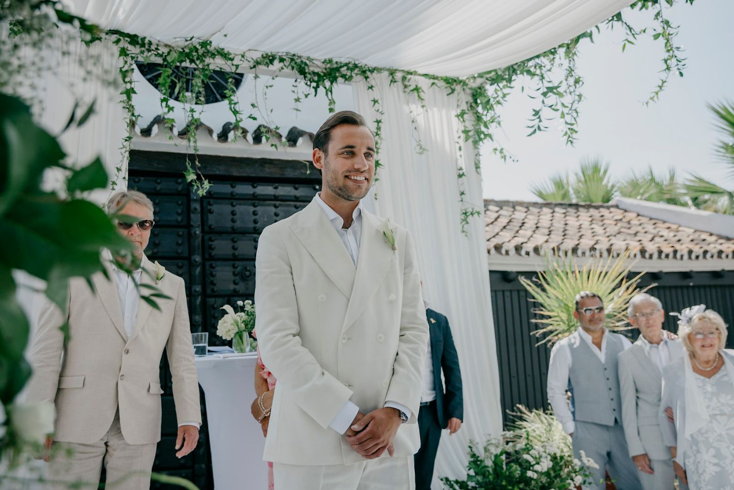 Outdoor wedding ceremony with white decor and greenery under a draped canopy.