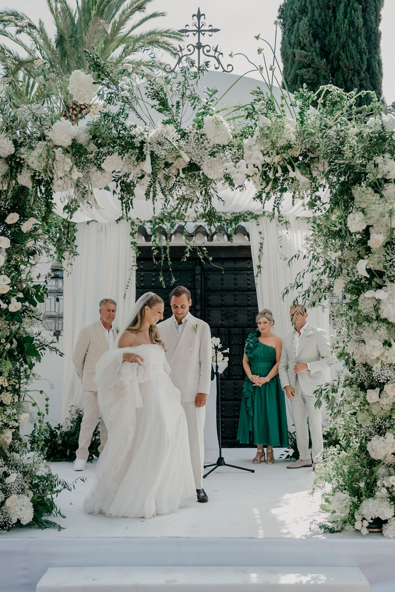 Wedding ceremony setup with white floral arch and decorative greenery against Mediterranean architecture.