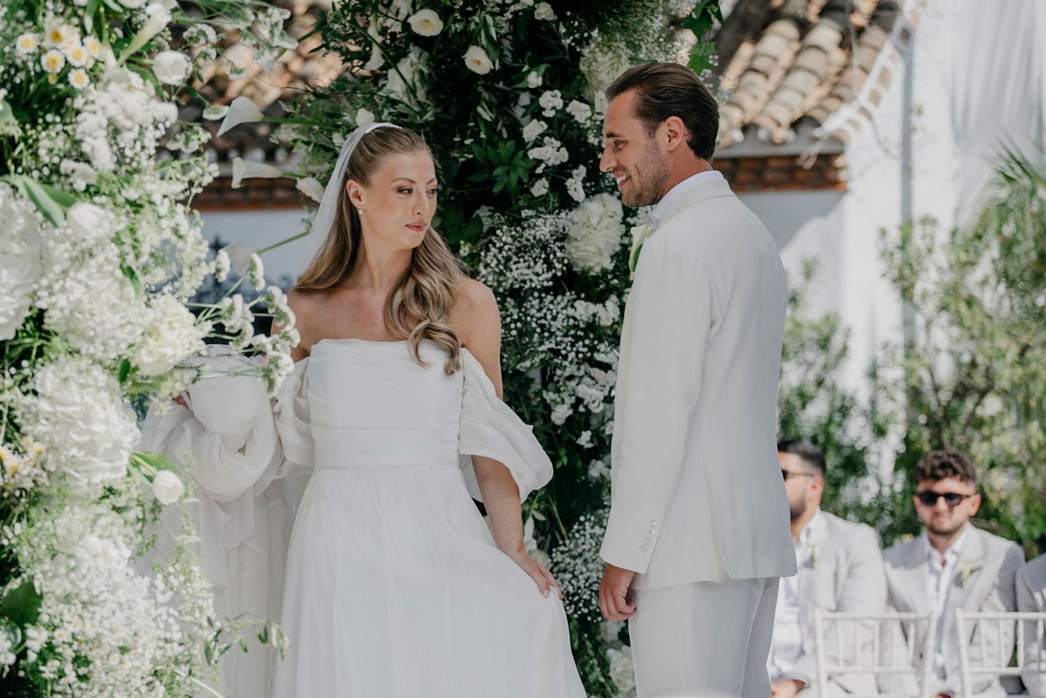 Wedding ceremony setup with white chairs and floral decorations under a Mediterranean-style architecture.