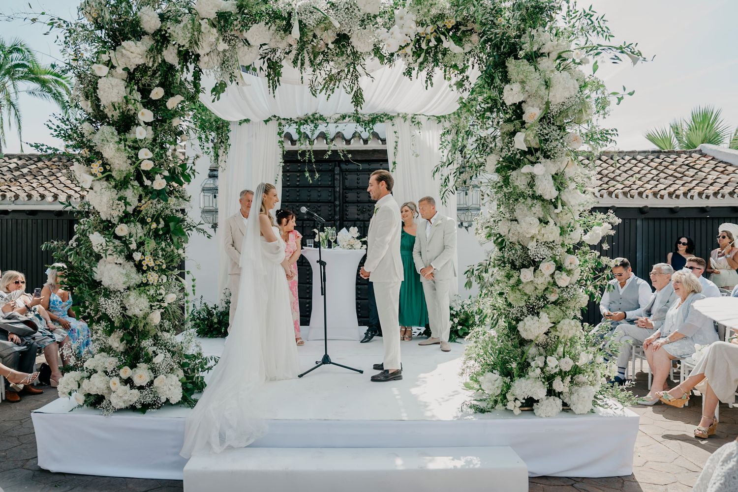 Elegant white floral arch and seating arrangement for an outdoor wedding ceremony.