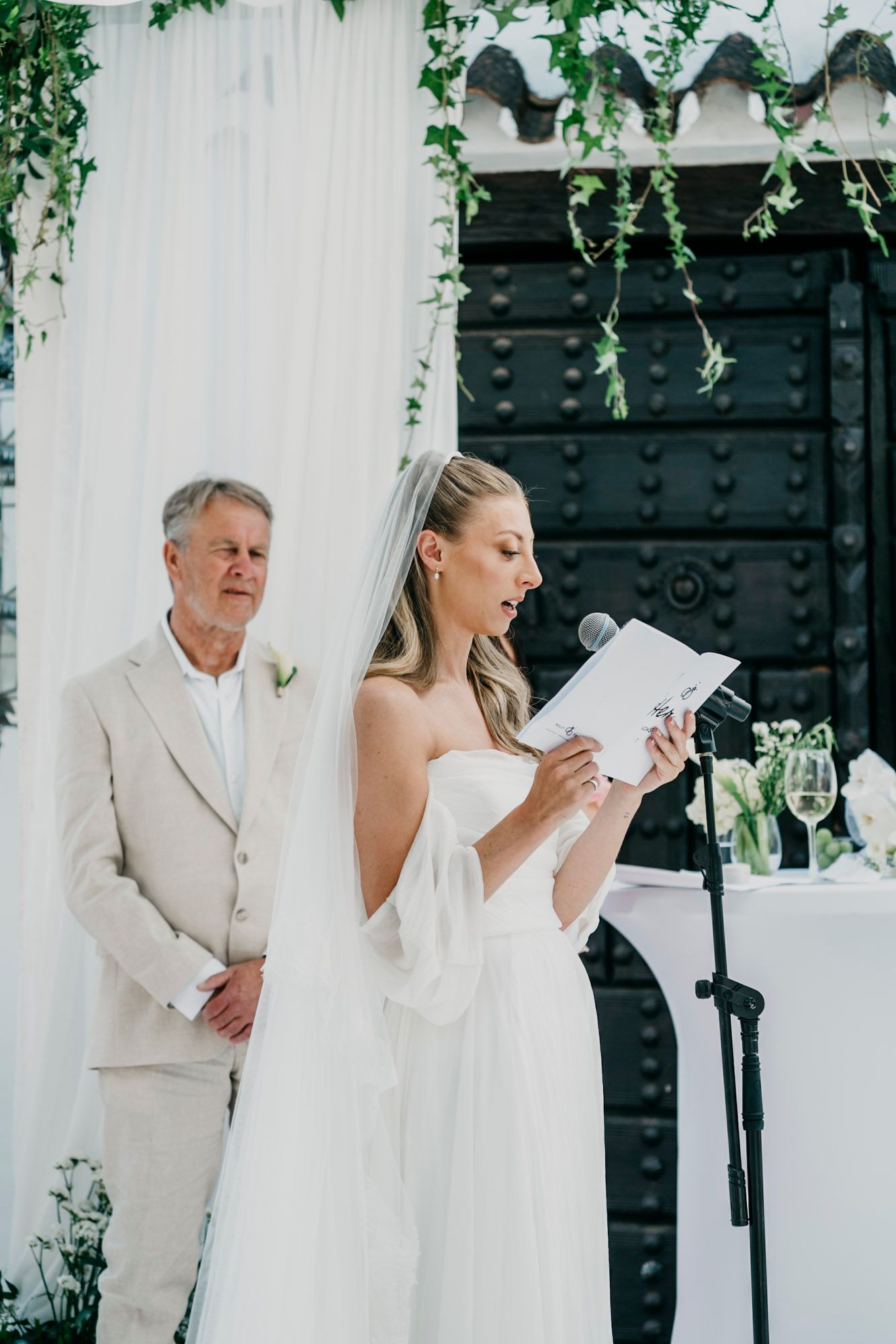 Wedding ceremony moment captured against a black door with hanging greenery and white decor.
