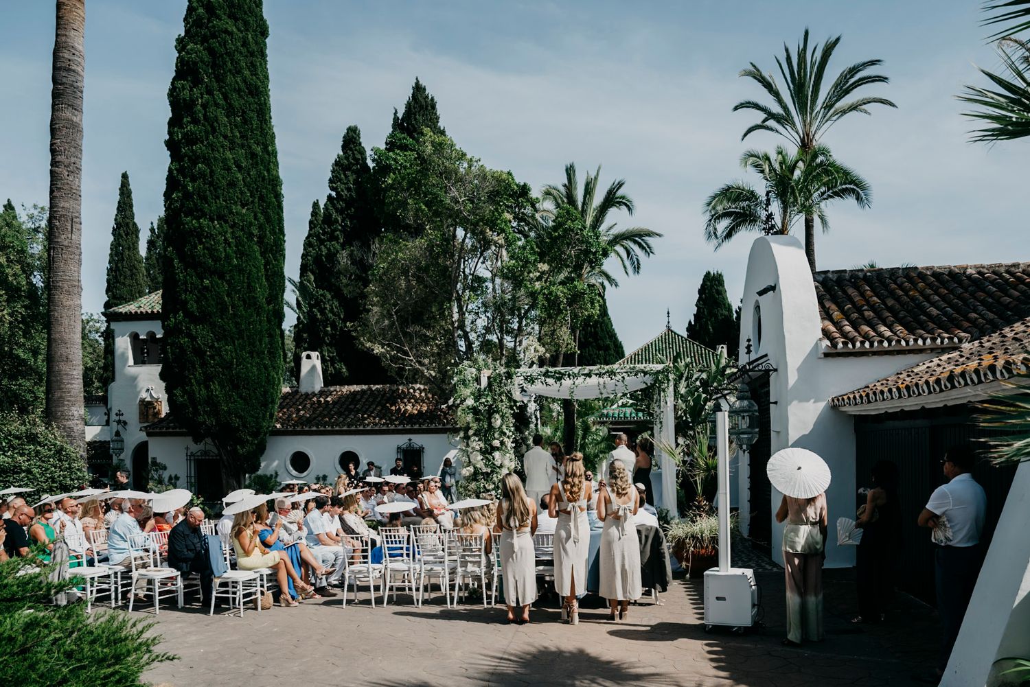 Outdoor Mediterranean wedding venue with tall cypress trees, palm trees and white chairs set up for ceremony.