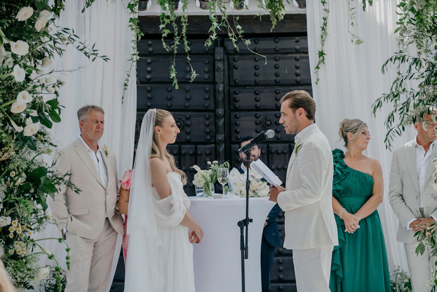 Wedding ceremony setup with white floral arrangements and greenery against a white wall.