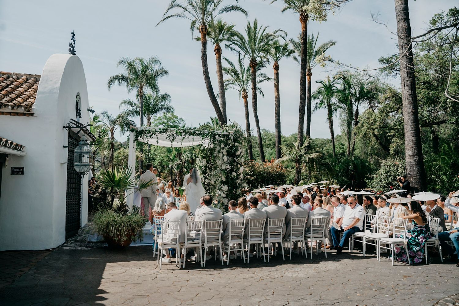 Outdoor wedding venue with rows of white chairs and palm trees against a Spanish colonial building.