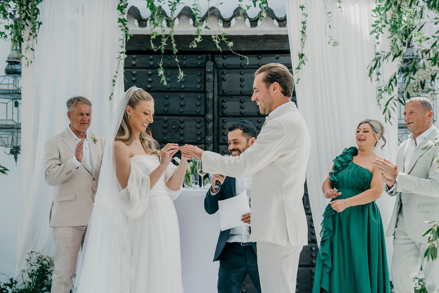 A wedding ceremony takes place outdoors with decorative greenery and attendants in green and white attire.
