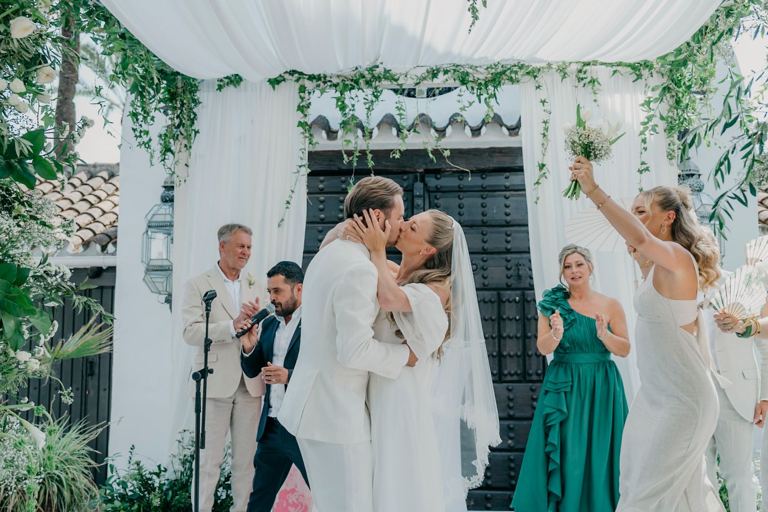 Wedding party celebrates under white drapes and hanging greenery at an outdoor reception.