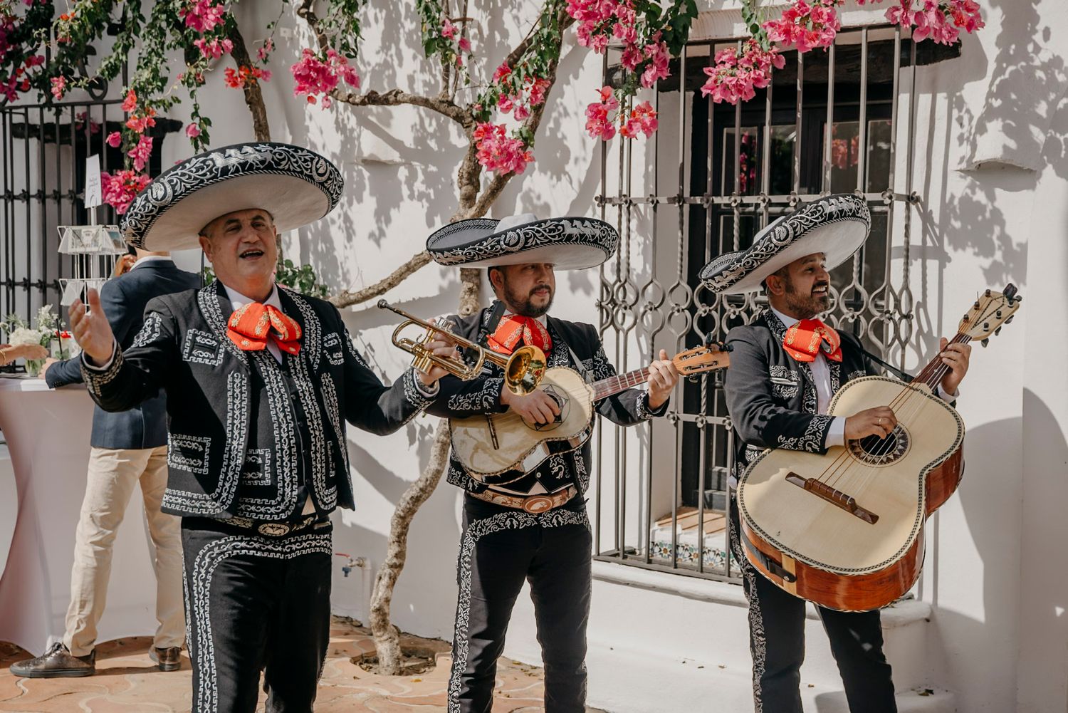 Mariachi band performers in traditional attire playing instruments against a white wall with pink flowers.