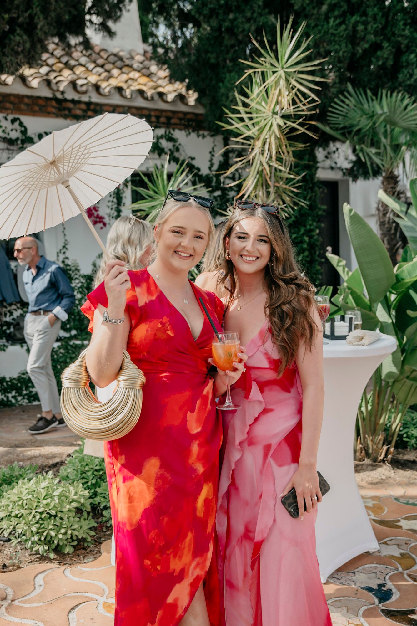 Garden party guests in bright red and pink dresses holding decorative parasols.