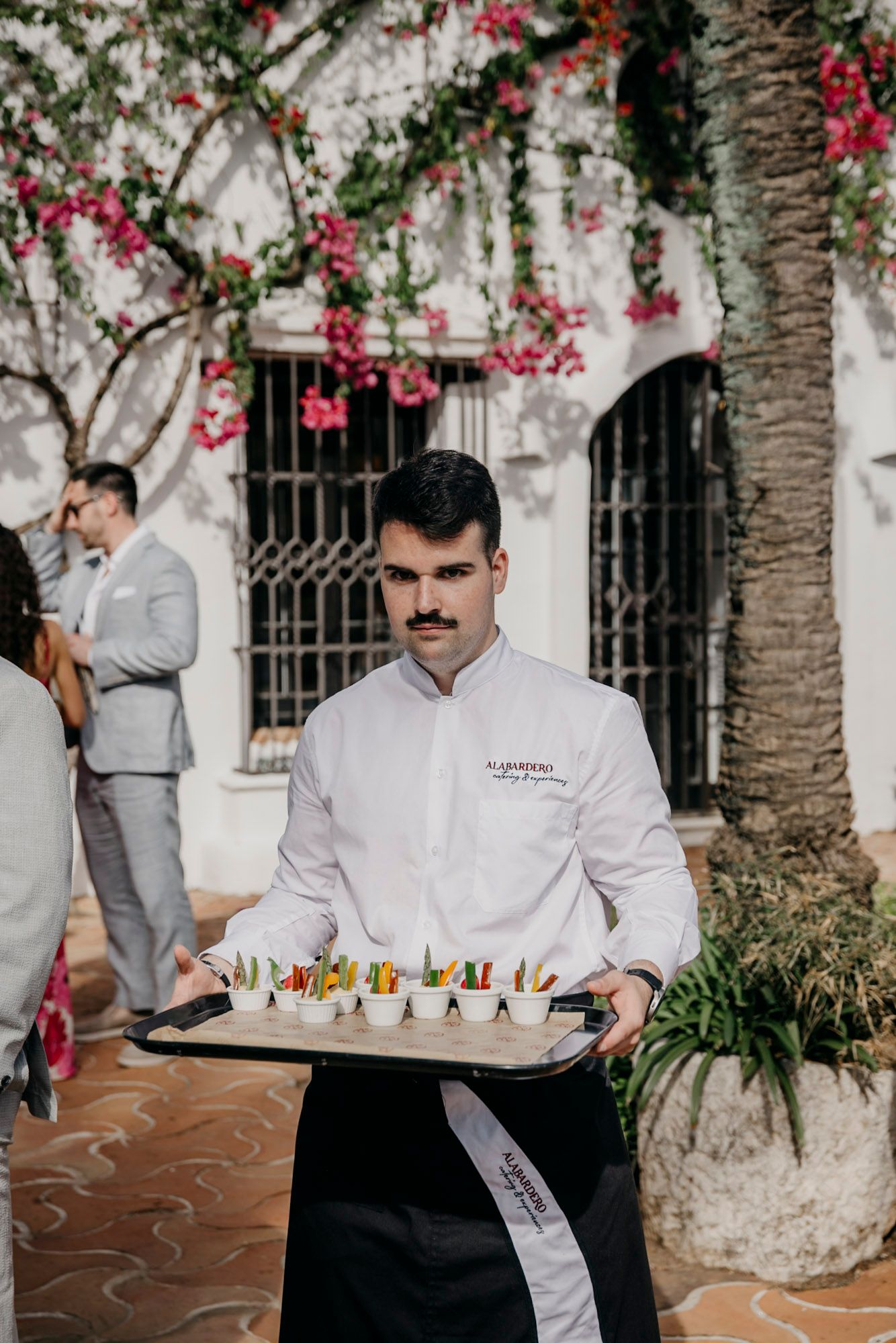Catering staff in white uniforms serving appetizers on trays at an outdoor event.