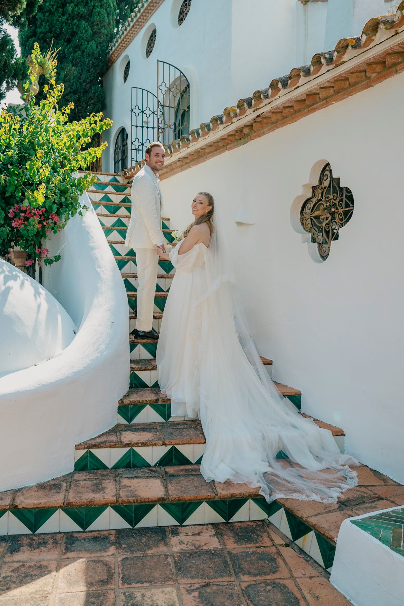 Bride in flowing white gown ascends decorative tiled steps at Mediterranean venue.