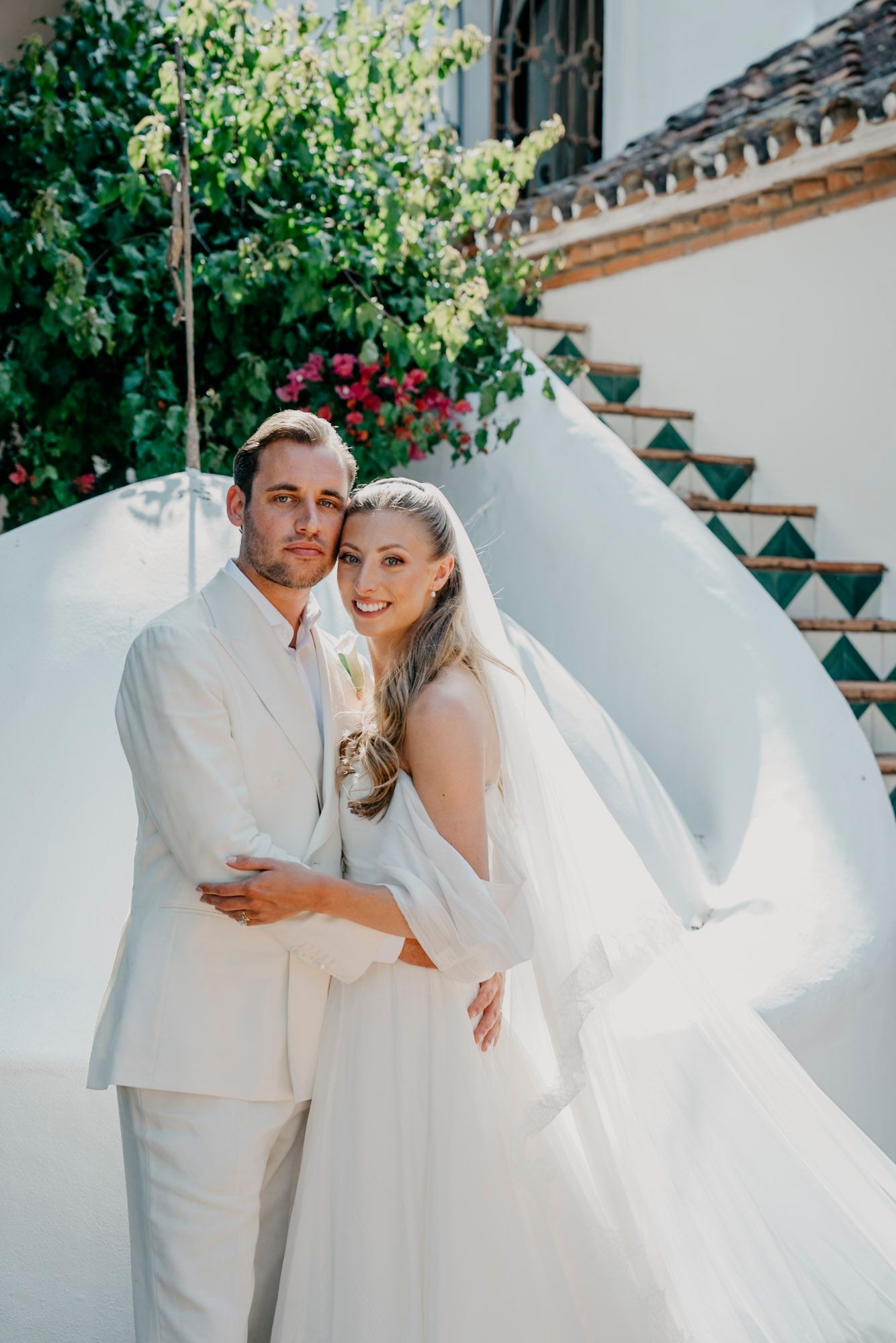 Newlyweds sharing romantic moment in white attire against garden wall.
