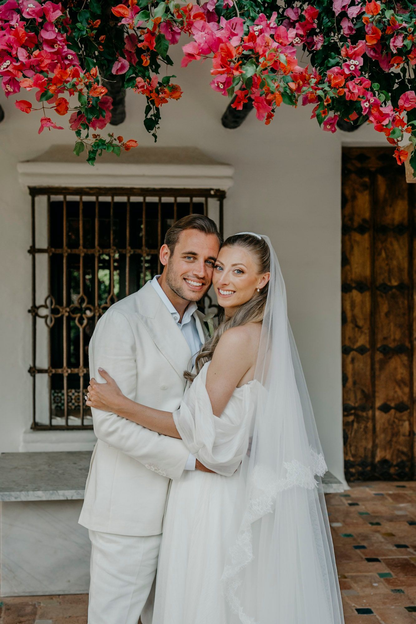 Wedding couple in all-white attire posing under vibrant pink bougainvillea flowers.