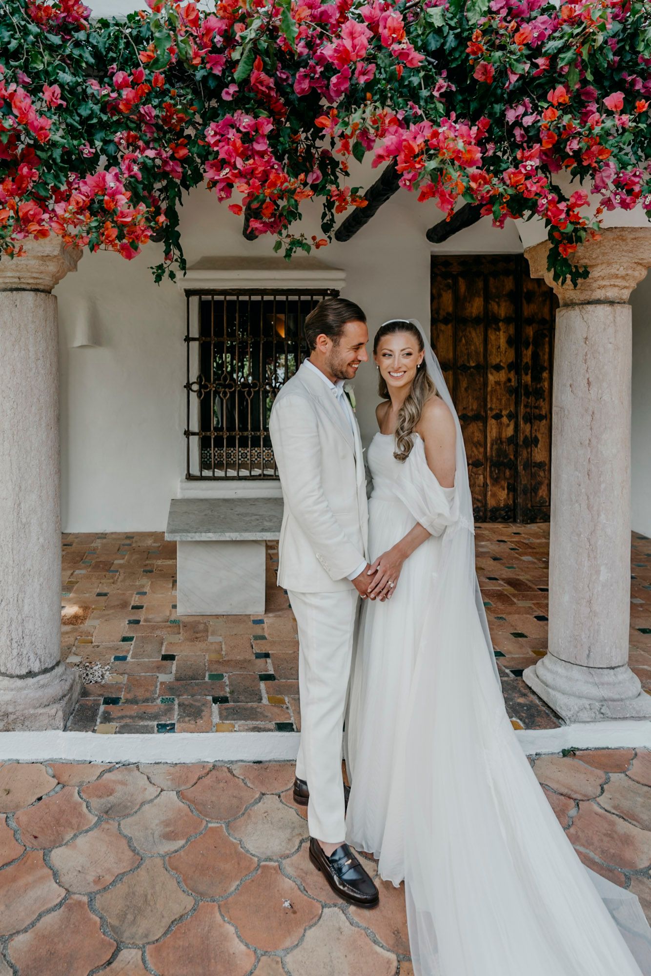 Newlyweds share intimate moments under vibrant pink bougainvillea flowers and white columns.