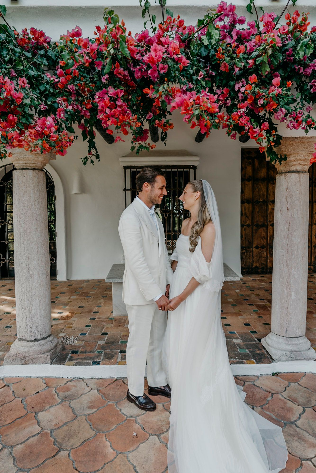 A couple in white stands under a colonnade covered with vibrant red bougainvillea flowers.