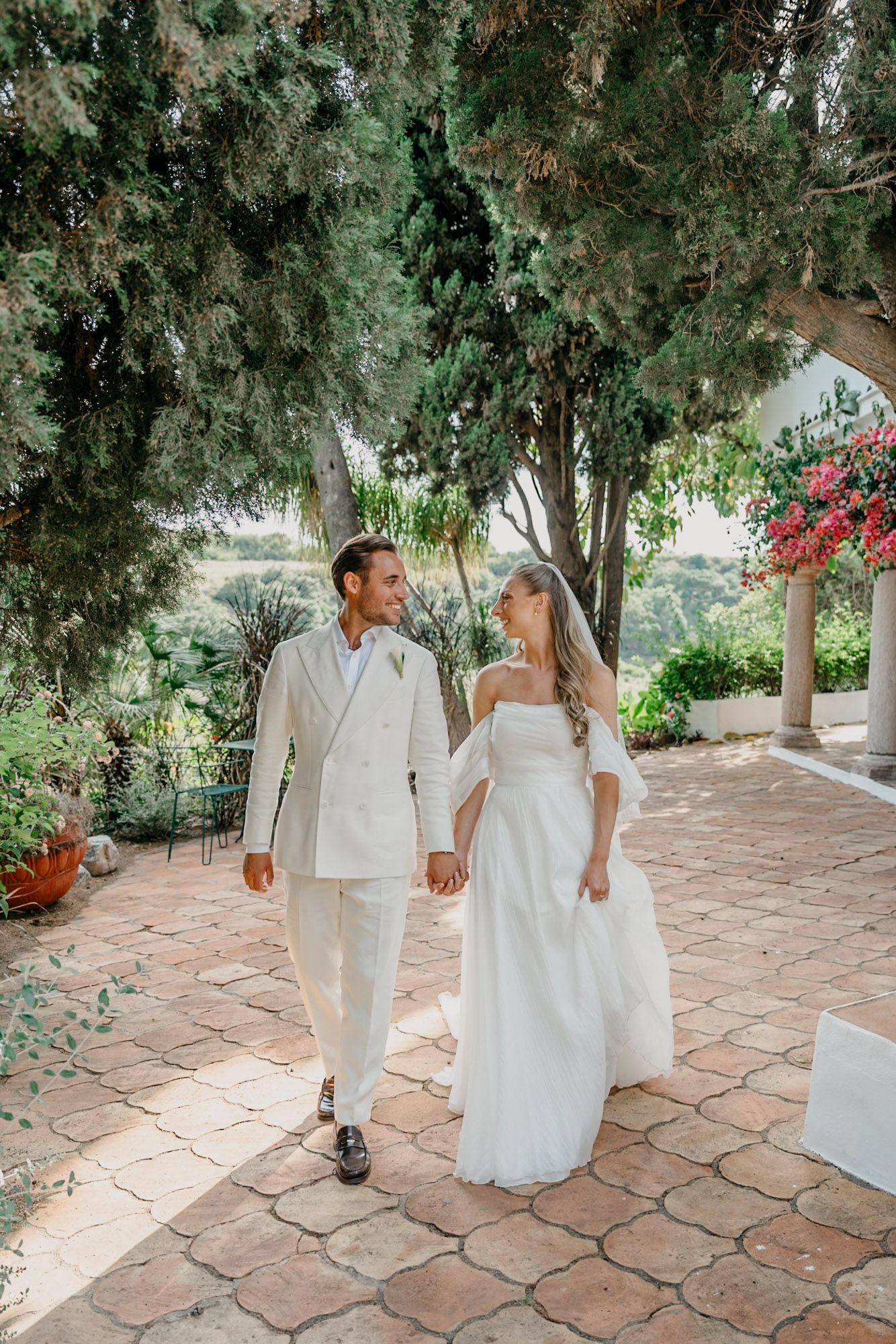 Newlyweds walking together on terracotta tiles surrounded by lush greenery and red flowers.