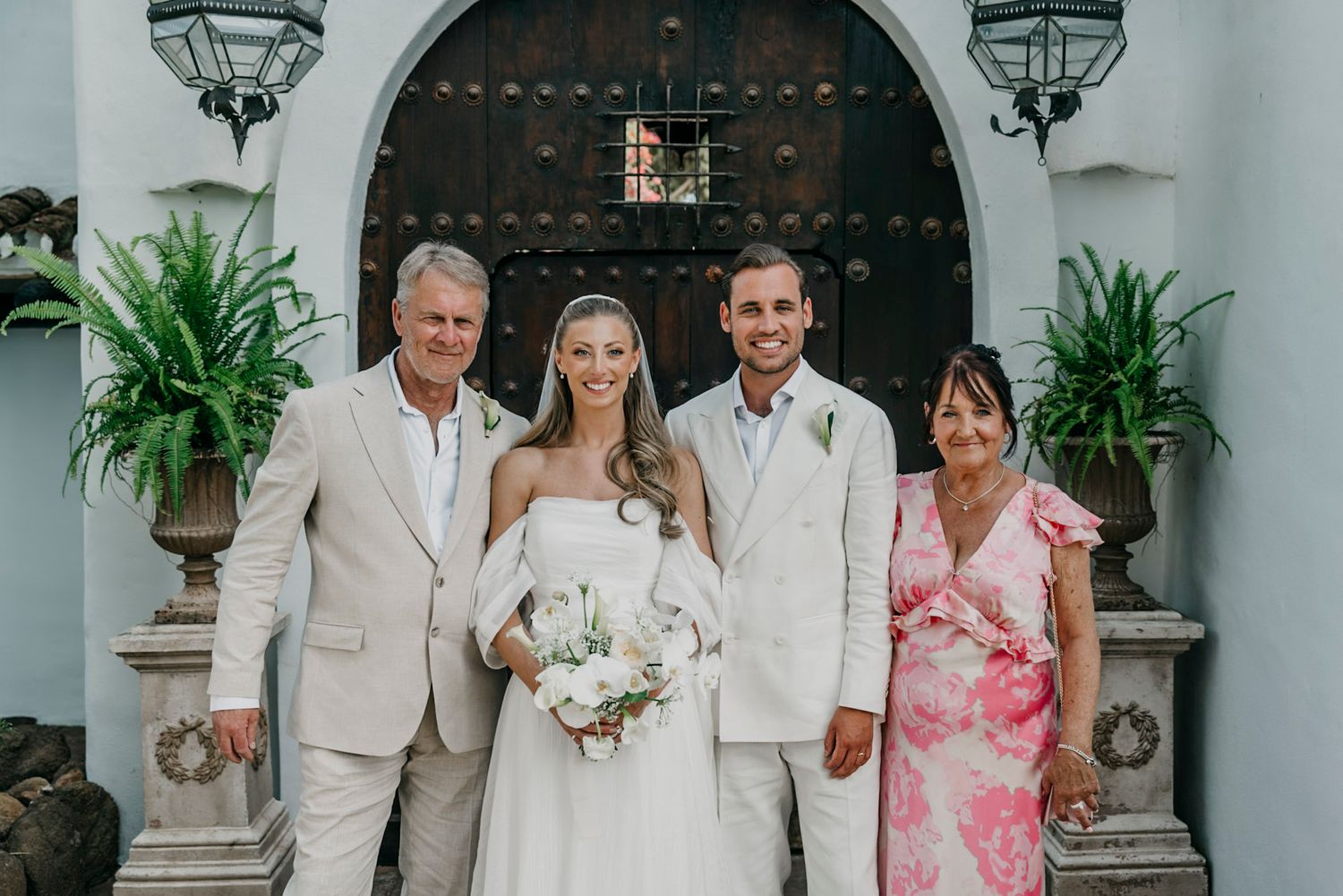 Wedding party in white and floral attire gathered in front of a Spanish colonial archway.