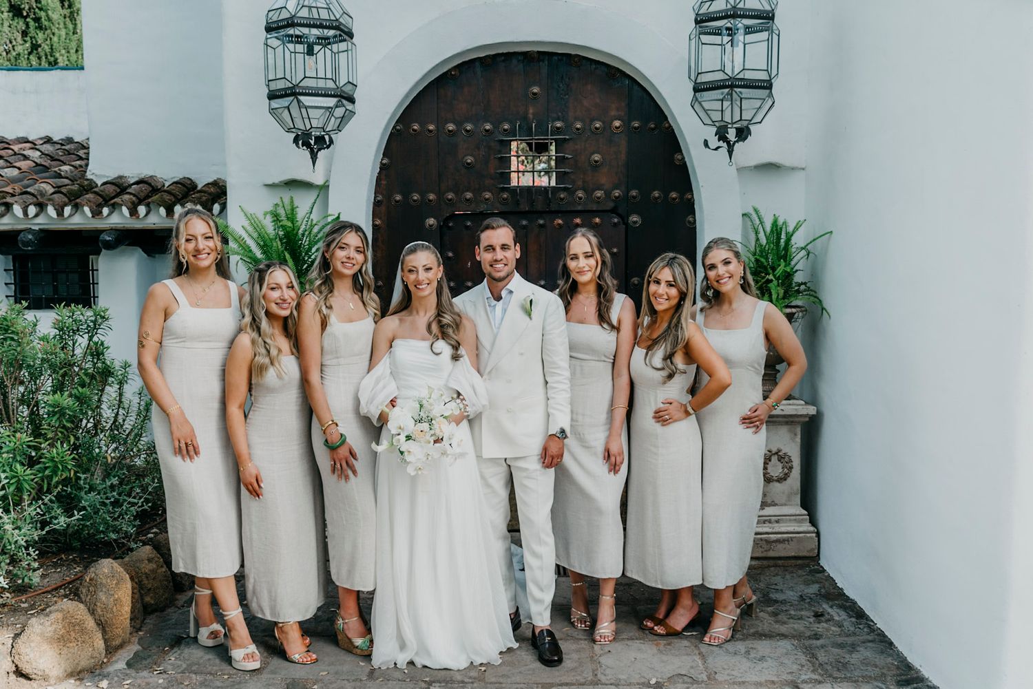 Wedding party posing for formal photos in front of a white Spanish colonial building.