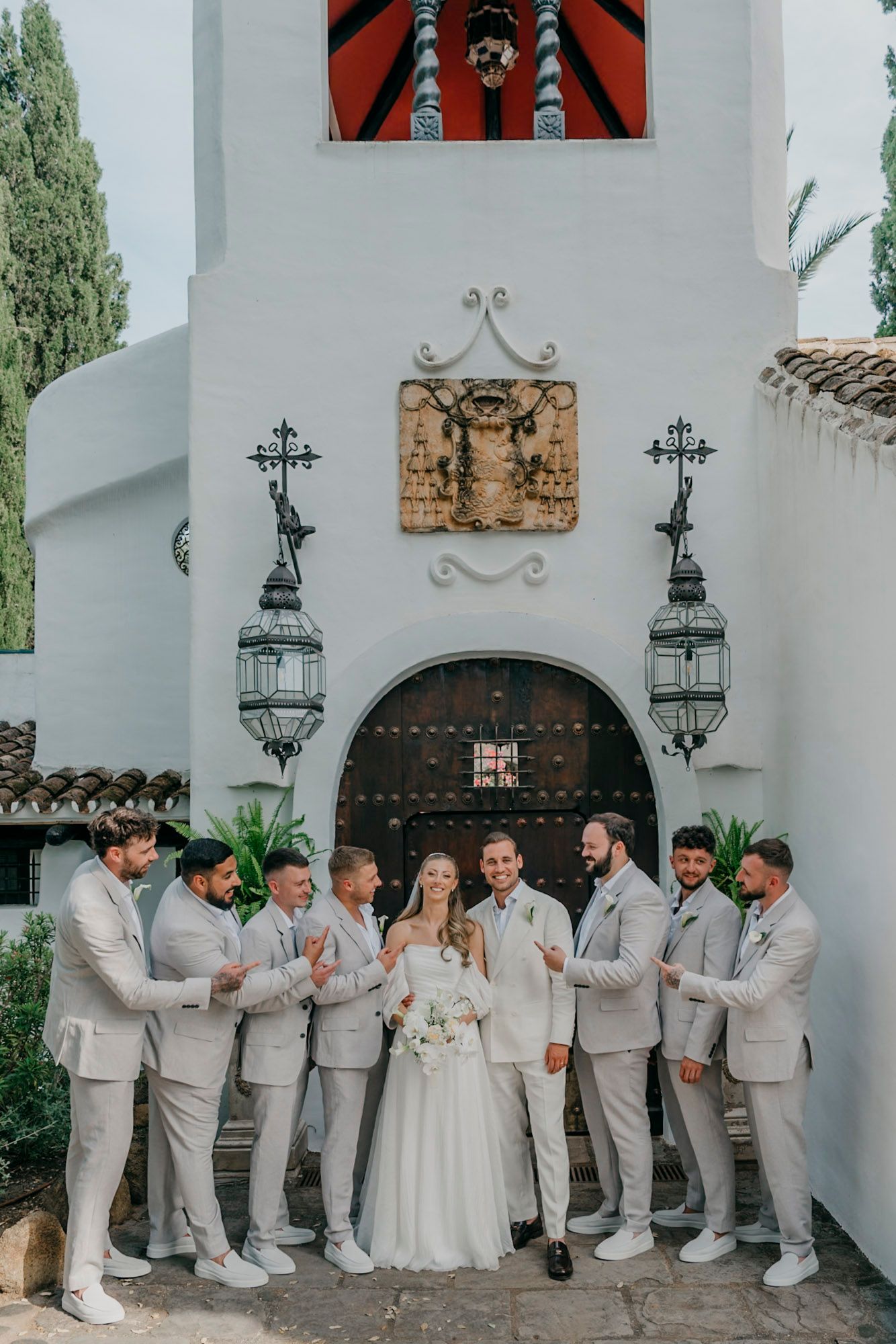 Wedding party in light gray suits poses against white Spanish-style church exterior.