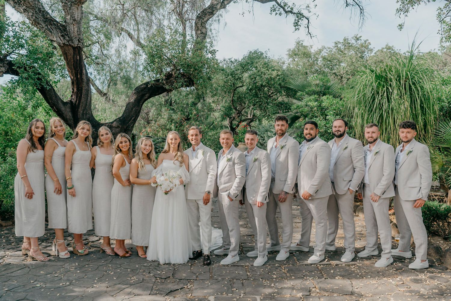 Wedding party in light gray attire poses together under trees in an outdoor garden setting.