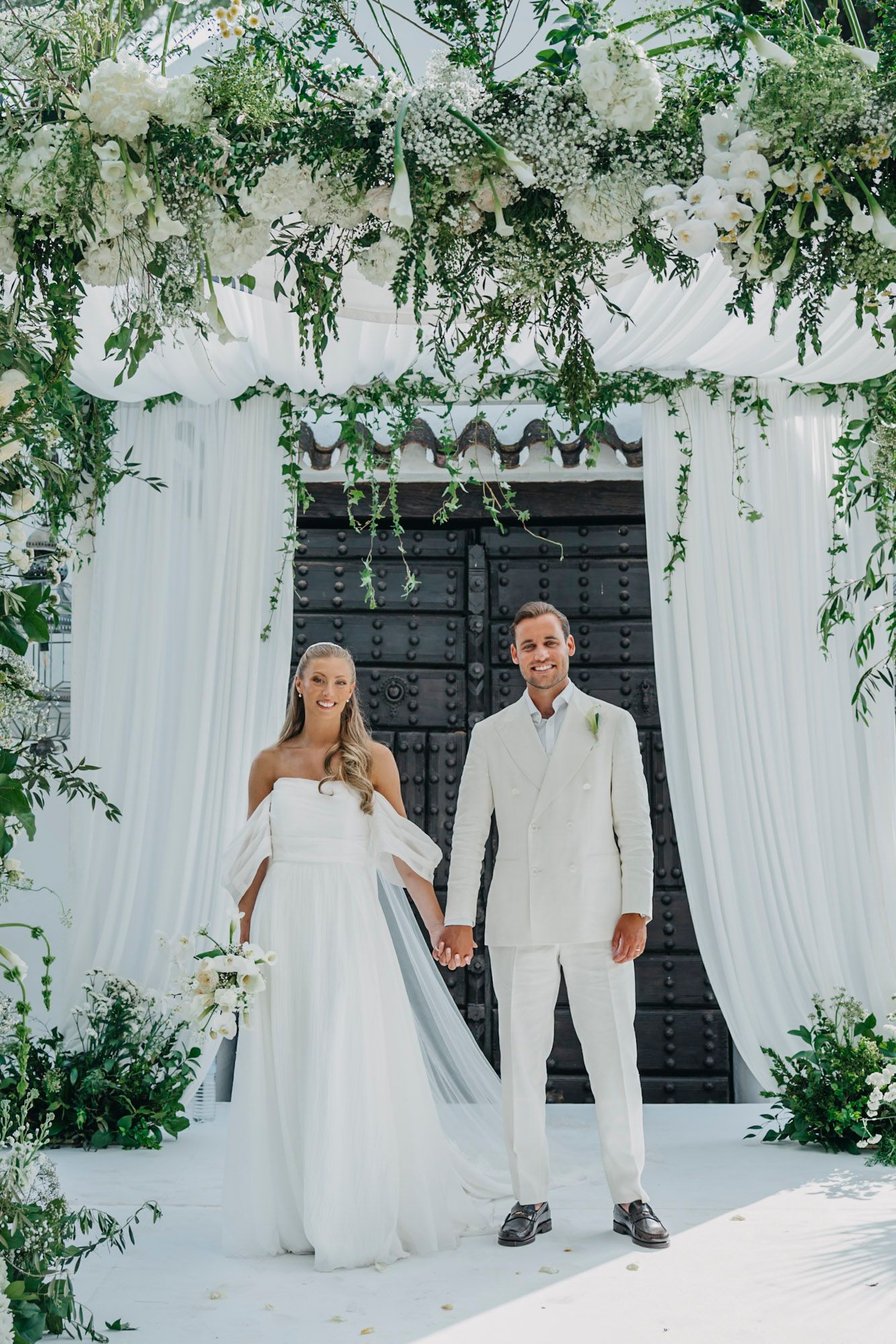 Wedding ceremony setup with floral arch and white draping against black door.