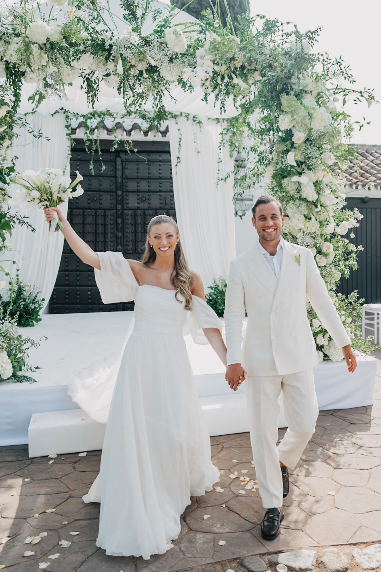Joyful couple celebrating under floral arch at outdoor wedding ceremony.