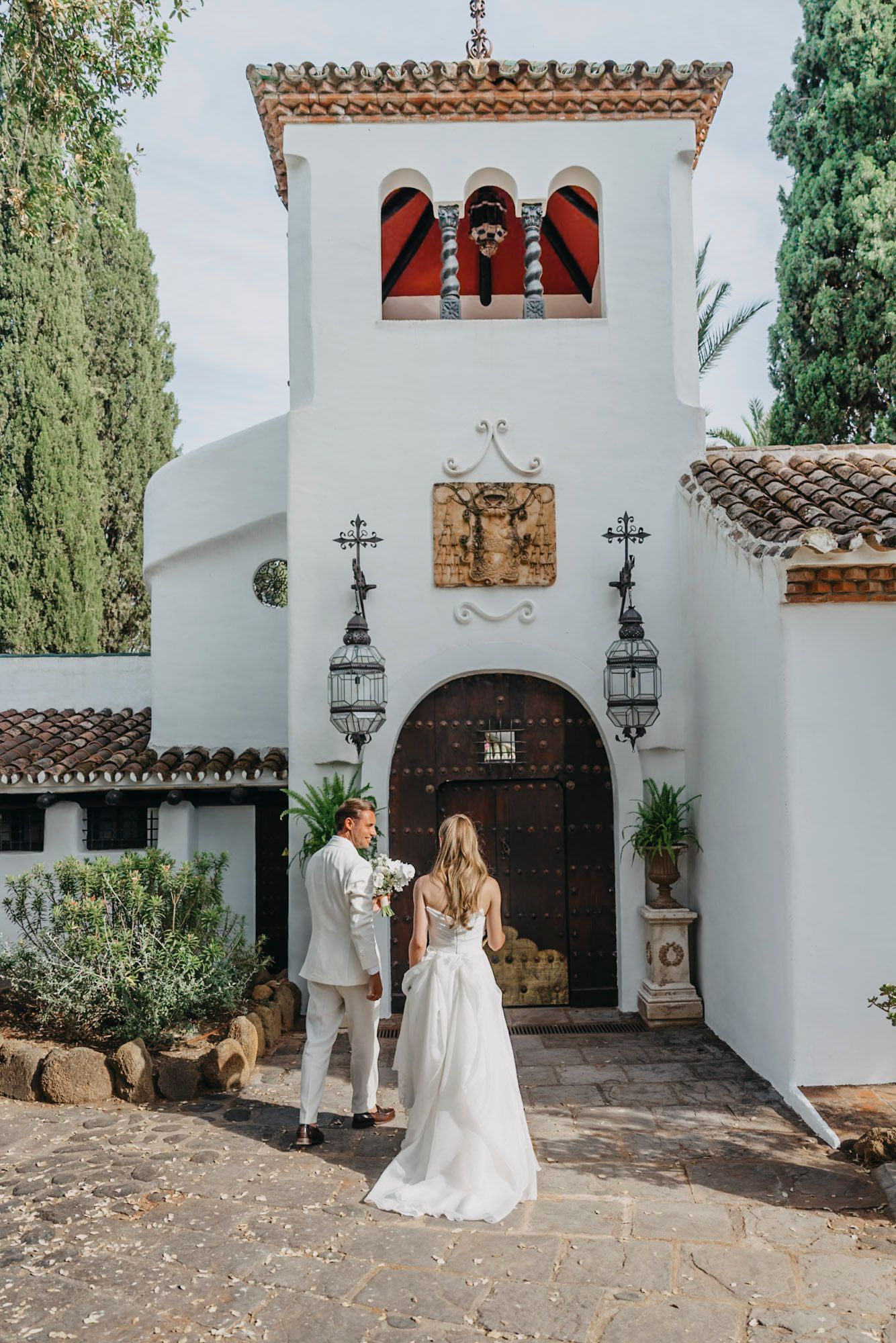 Spanish colonial church entrance with white walls, wooden doors, and ornate lanterns during a wedding ceremony.