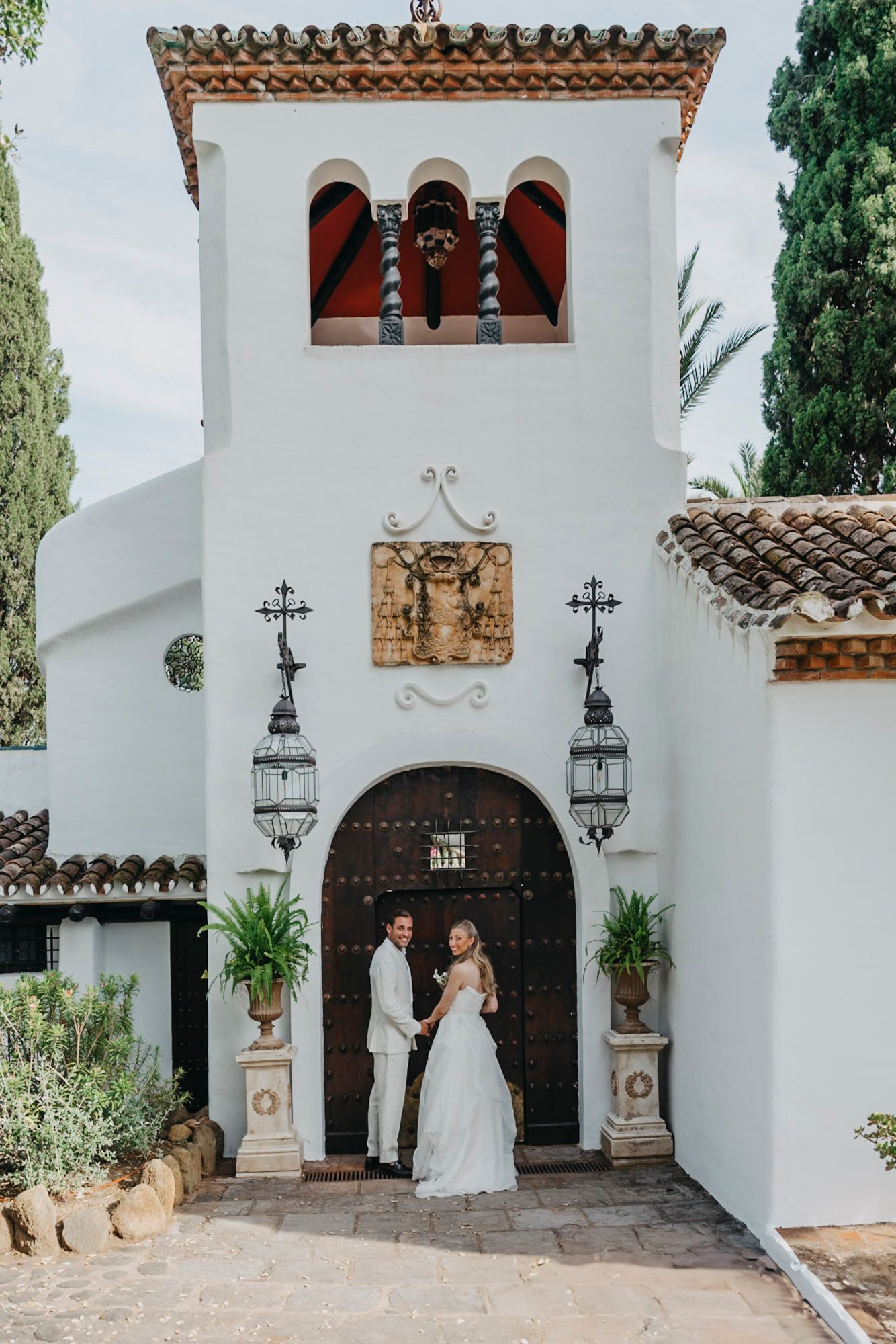 Spanish-style white church entrance with ornate bell tower and decorative lanterns framing an arched doorway.