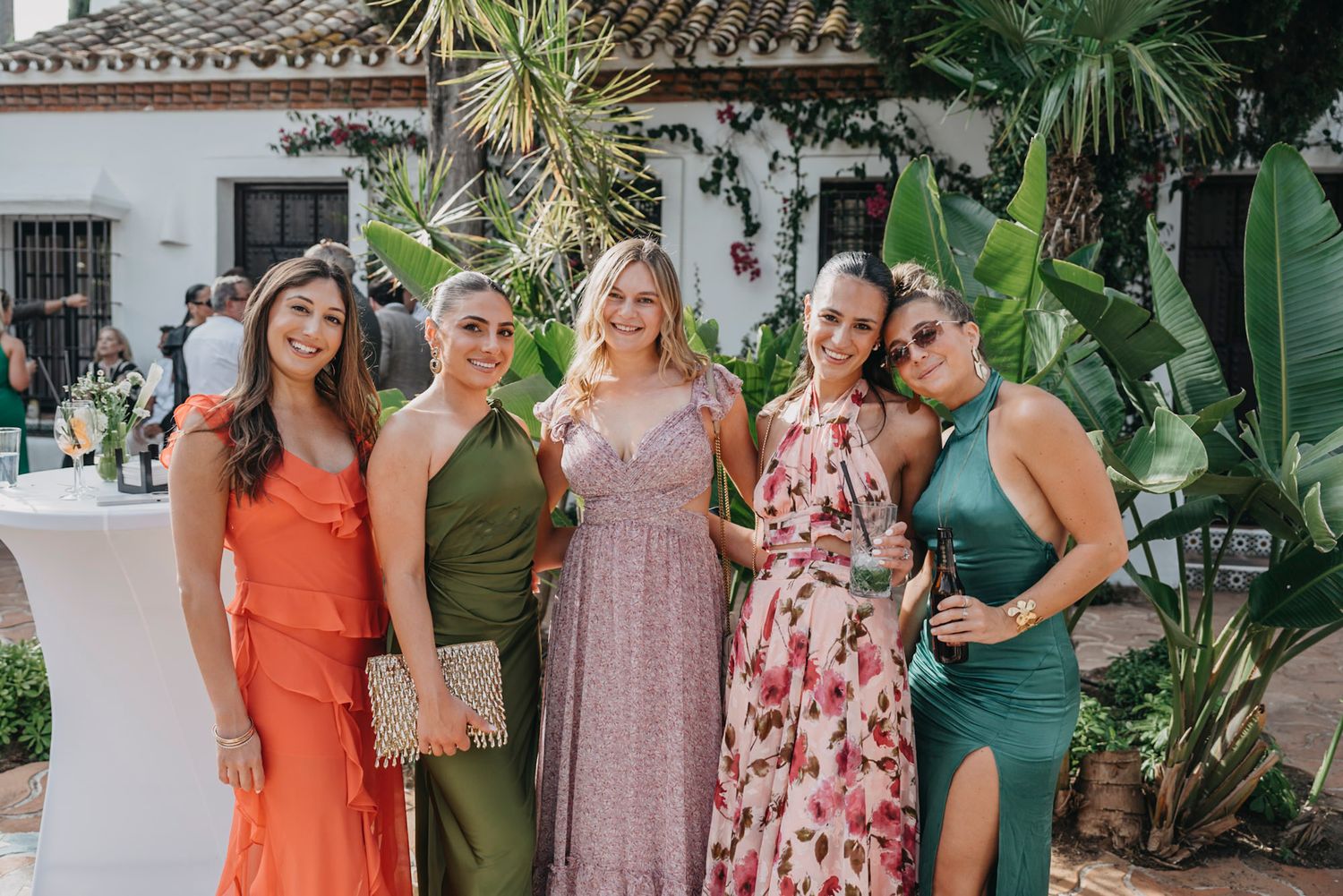 Wedding guests in colorful summer dresses gather outdoors at an elegant venue with tropical landscaping.