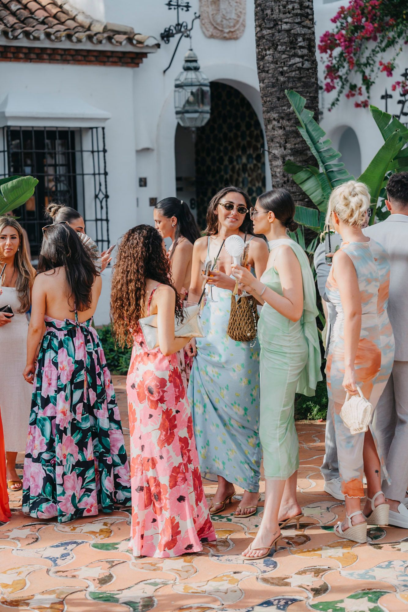 A group of guests in colorful floral dresses gather at an outdoor Spanish-style venue with white walls and palm trees.