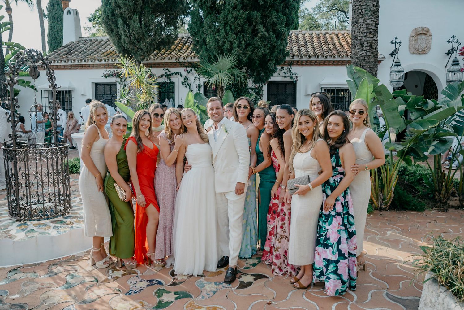 Wedding guests in colorful dresses and suits gathered for a group photo in a garden setting.
