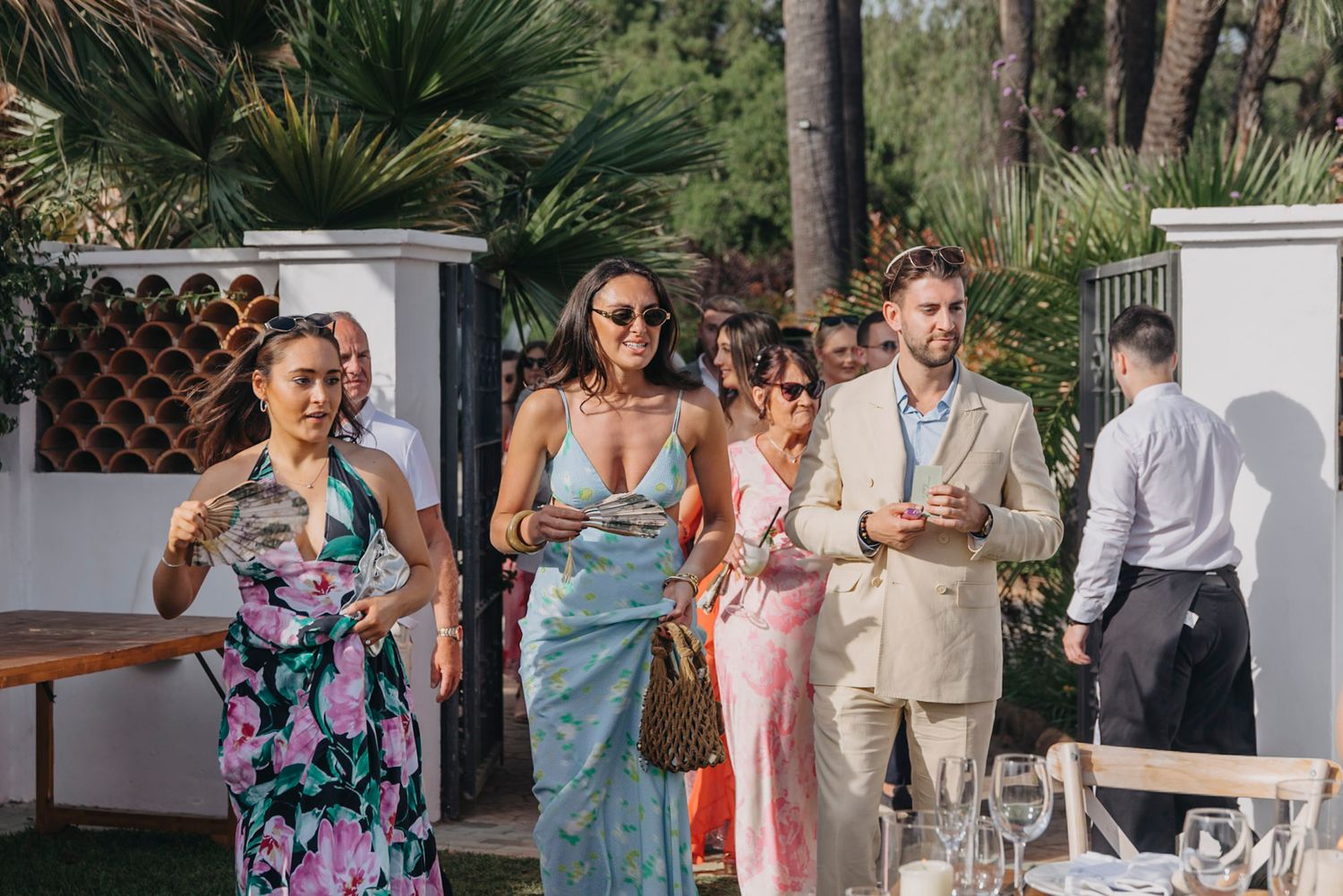 Wedding guests in formal attire enjoy drinks and conversation during an outdoor reception at a tropical venue.