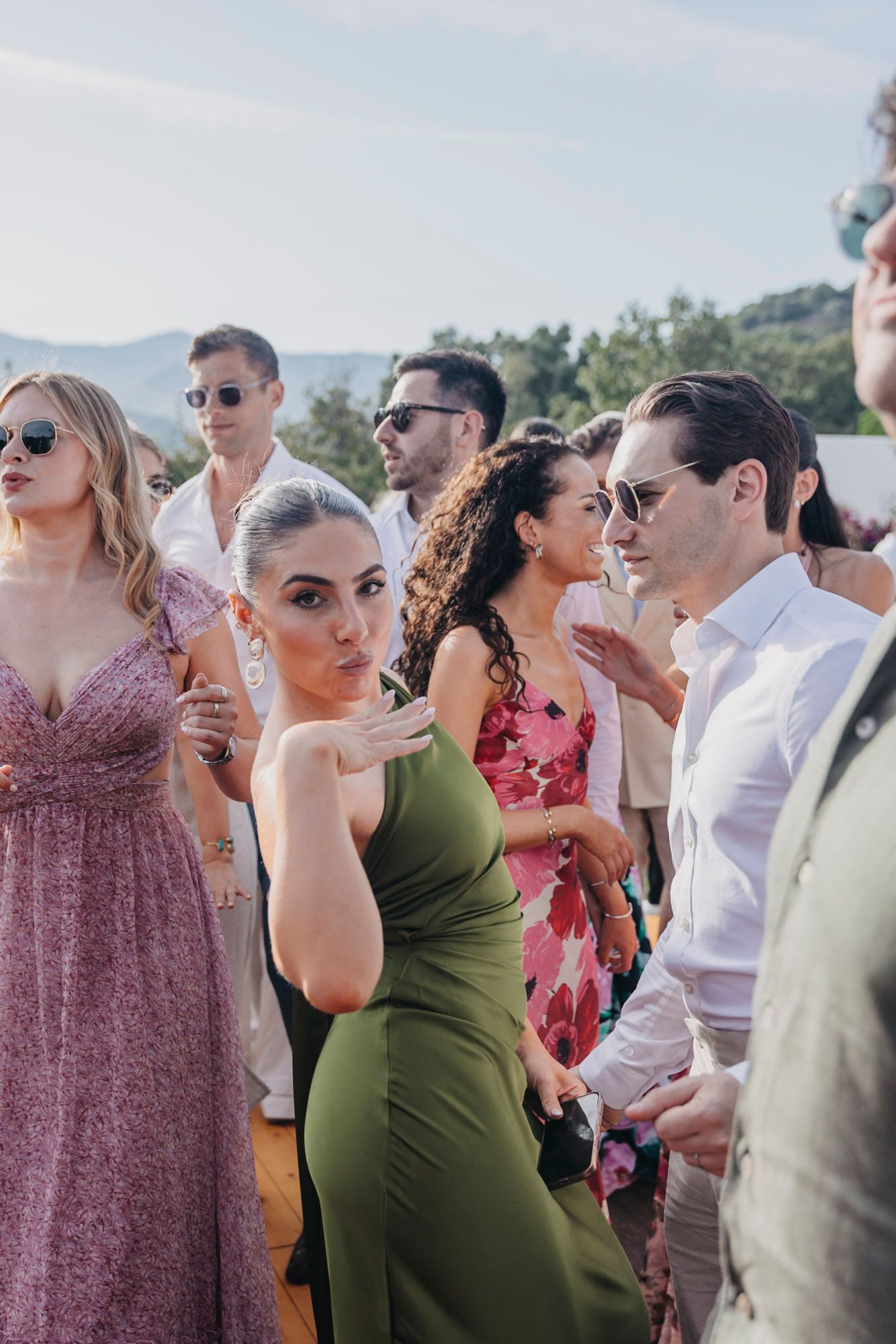 Wedding guests mingle at an outdoor reception wearing summer attire and sunglasses.