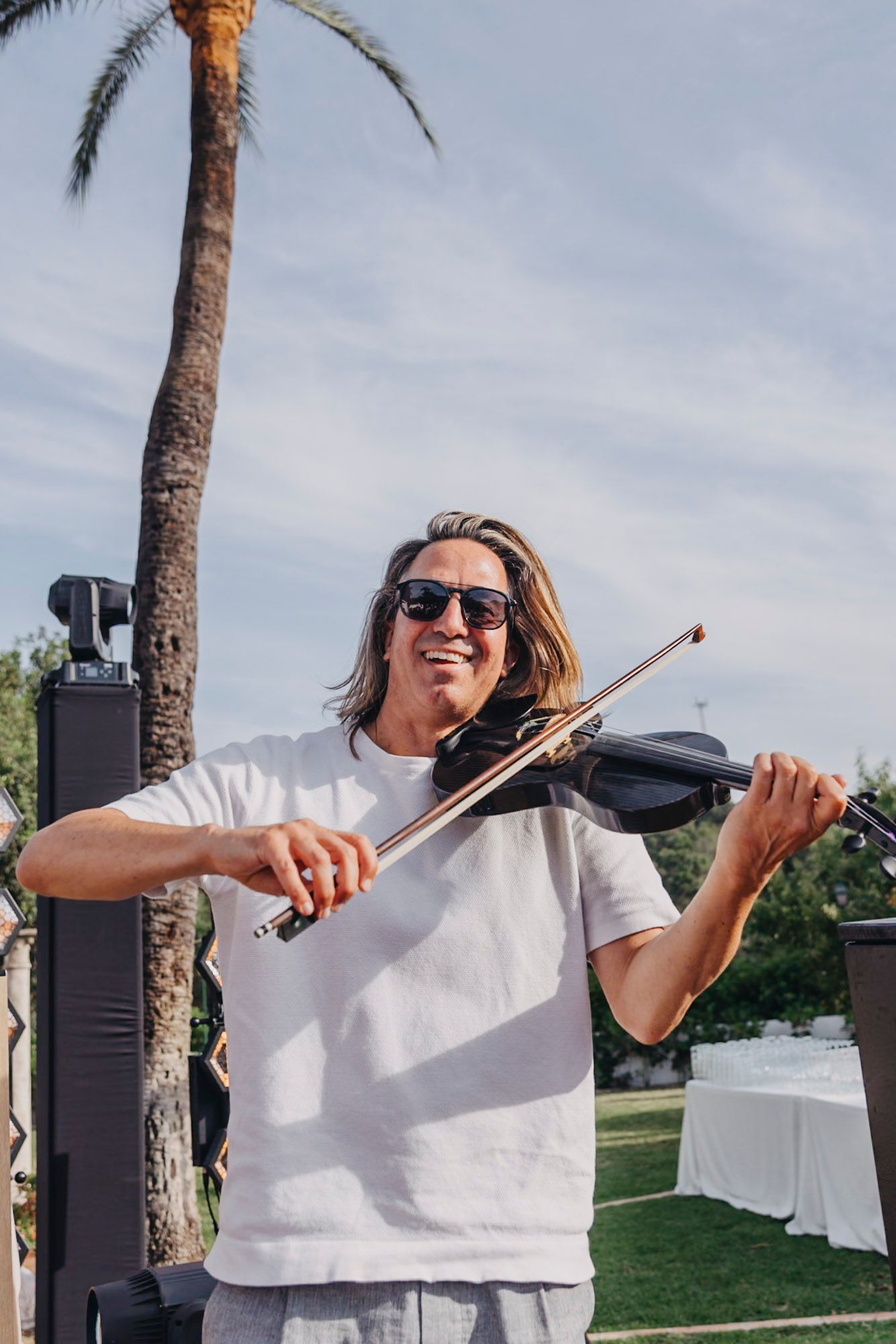 Person in white shirt plays violin energetically outdoors with palm trees in background.