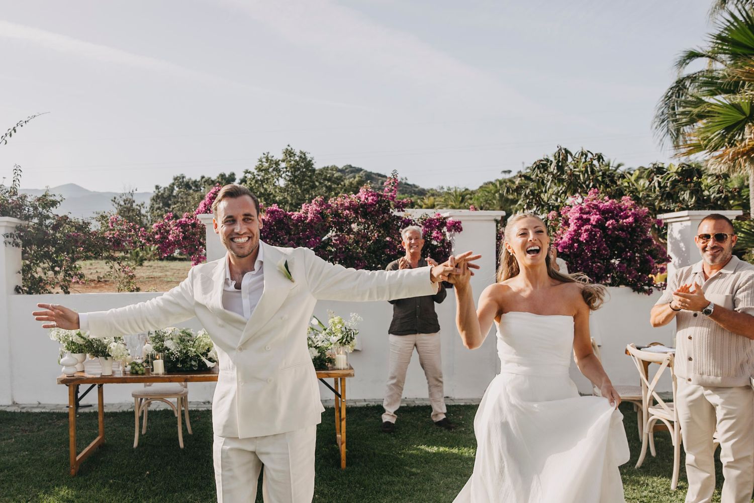 Wedding party poses for photos in an elegant outdoor garden setting with white attire.