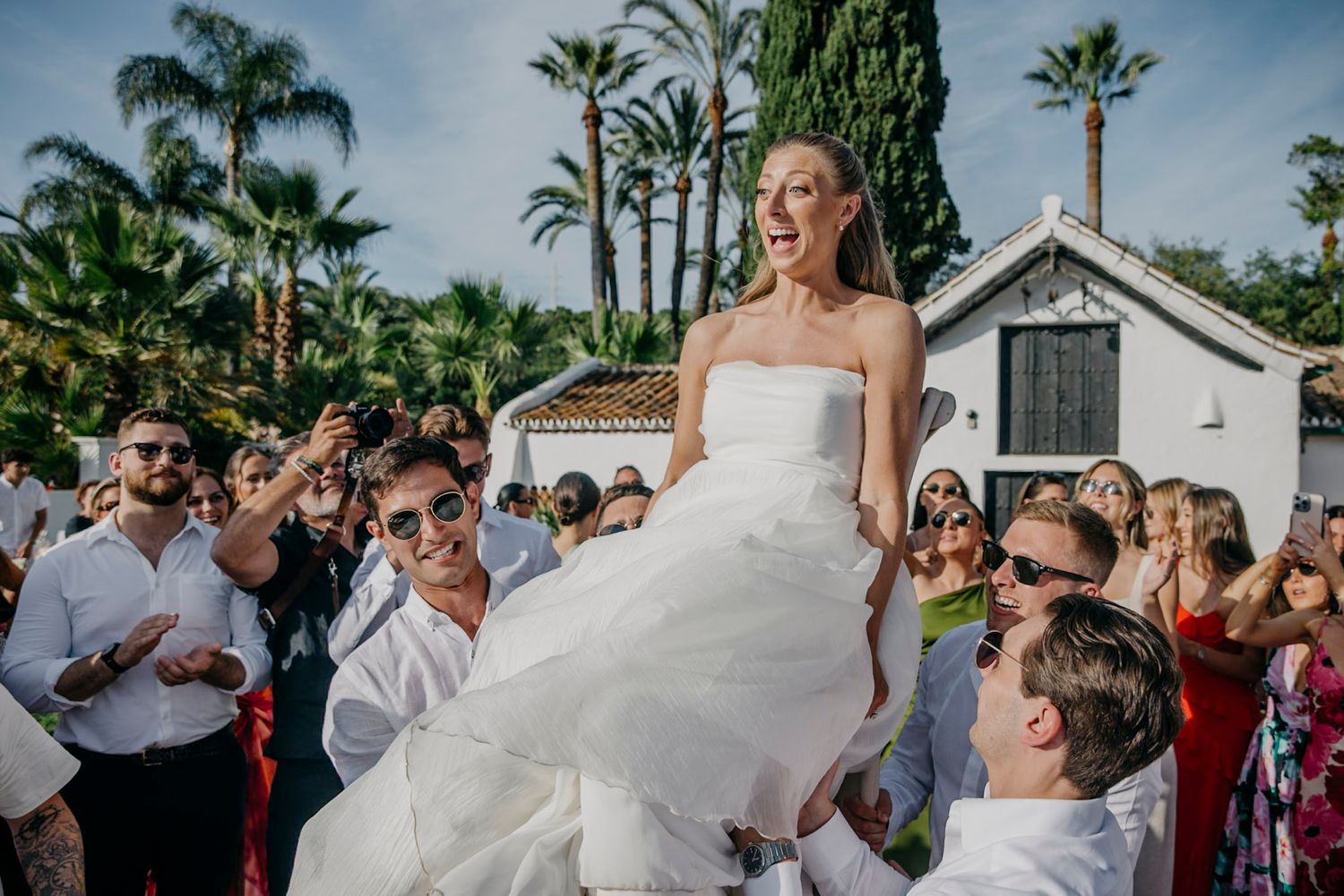 Wedding celebration with guests dancing and jumping joyfully outdoors under palm trees.