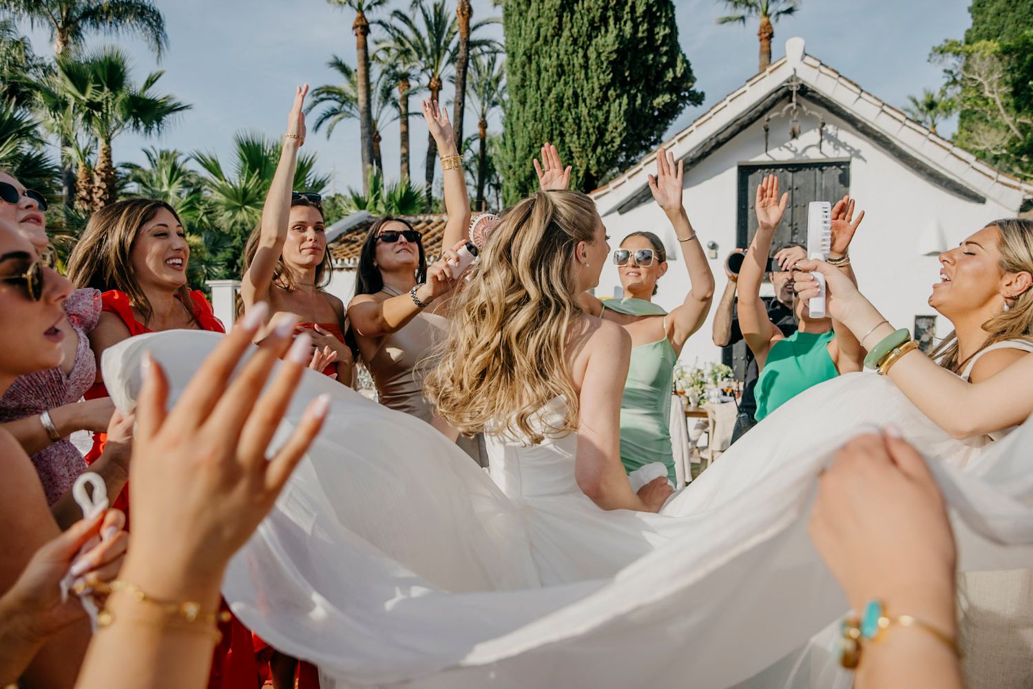Wedding guests celebrate and dance outdoors with a flowing white veil against a backdrop of palm trees.