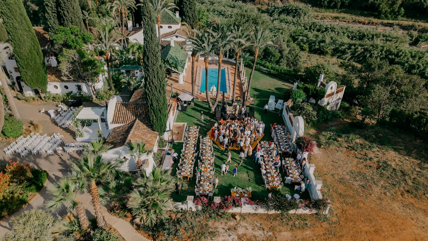 Aerial view of an elegant outdoor wedding ceremony with rows of white chairs in a lush garden setting.