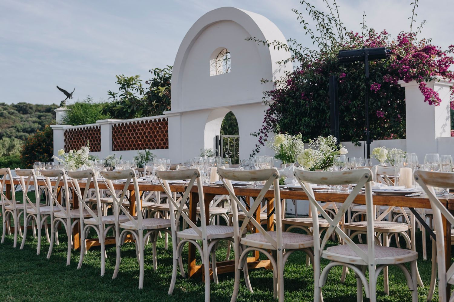 Rustic wooden cross-back chairs arranged for an outdoor wedding ceremony with white archway.