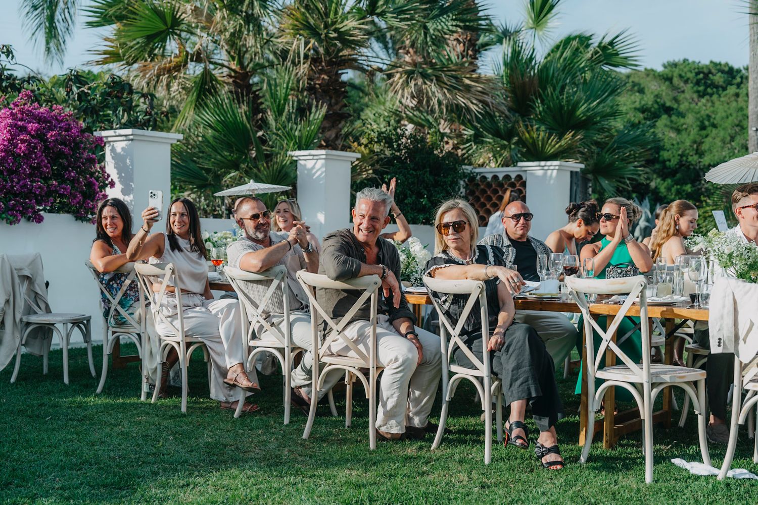 Wedding guests seated in white chairs at an outdoor tropical venue with palm trees and greenery.