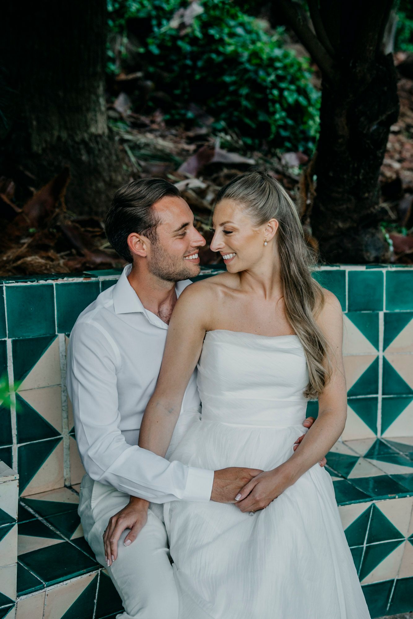 Romantic couple in white attire pose together against a green and teal geometric wall backdrop.