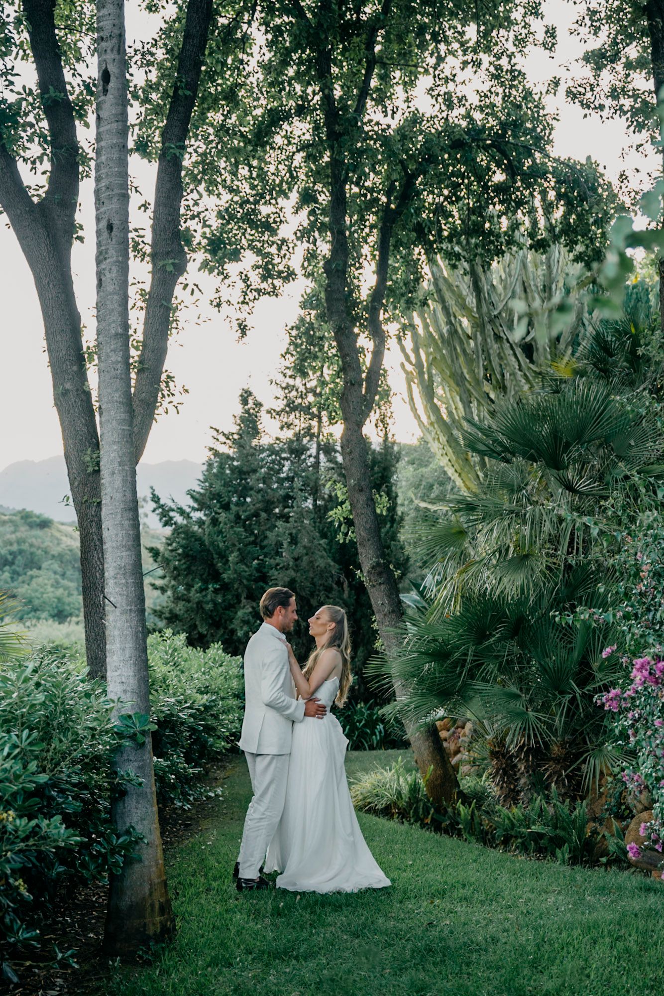 A romantic wedding couple shares intimate moments in a lush garden setting with palm trees and greenery.