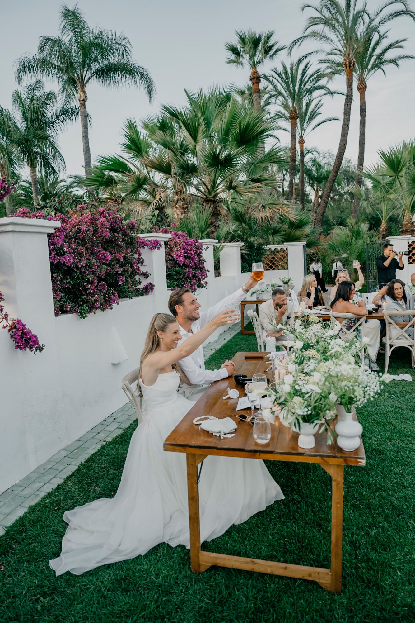 Wedding party enjoying drinks and toasts at an elegant outdoor garden reception with palm trees and bougainvillea.