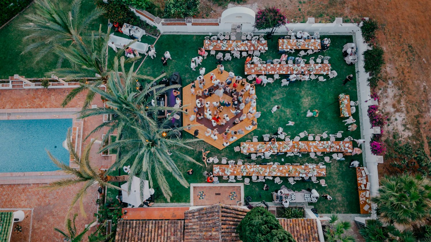 Aerial view of an outdoor wedding ceremony with guests seated in a circular pattern around a central gazebo on green lawn.
