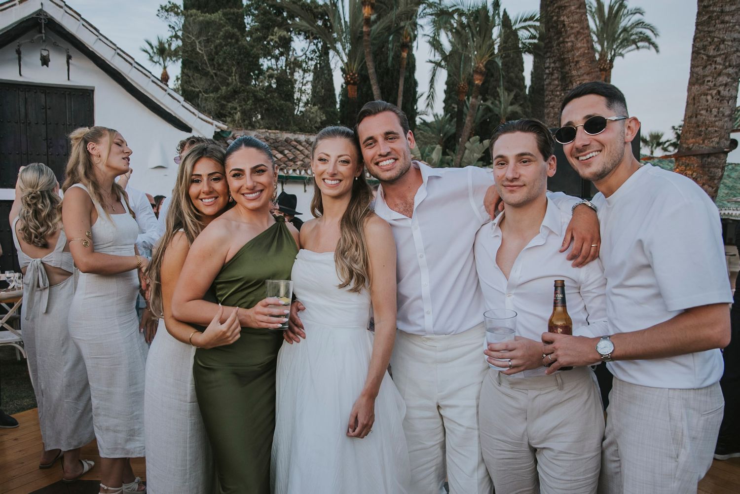 A group in coordinated white outfits poses together at an outdoor celebration with palm trees in the background.