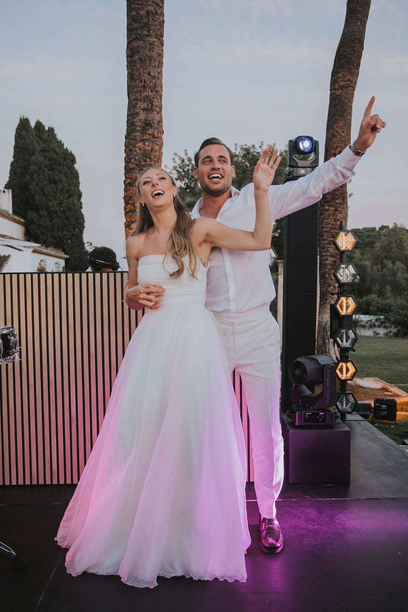 Newlyweds dancing joyfully at their wedding reception with purple lighting and palm trees in the background.