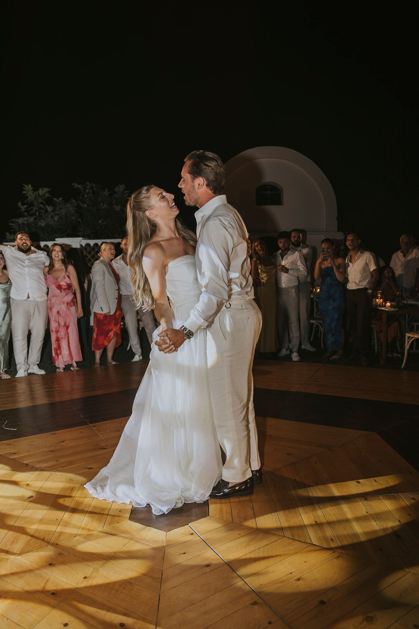 A couple shares a romantic dance on a wooden floor illuminated by spotlights during their wedding reception.