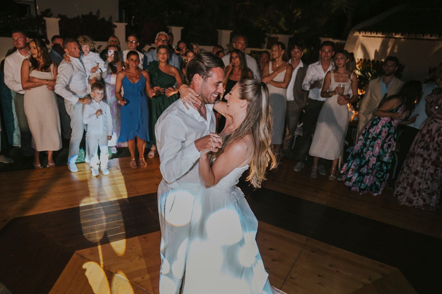 Wedding guests dance and celebrate on a dimly lit indoor dance floor during an evening reception.