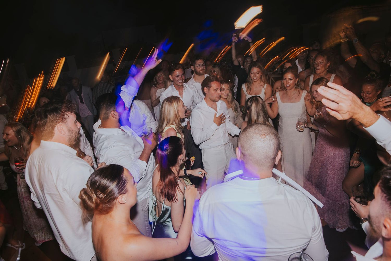 Wedding guests dancing and celebrating on a crowded dance floor with colorful lighting.