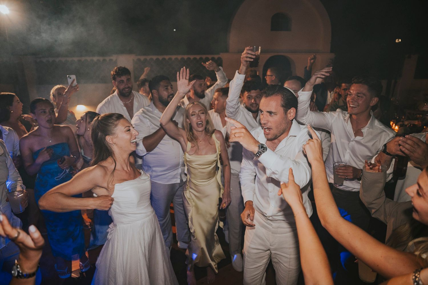 Wedding guests dance and celebrate together on a dark dance floor at a lively reception party.
