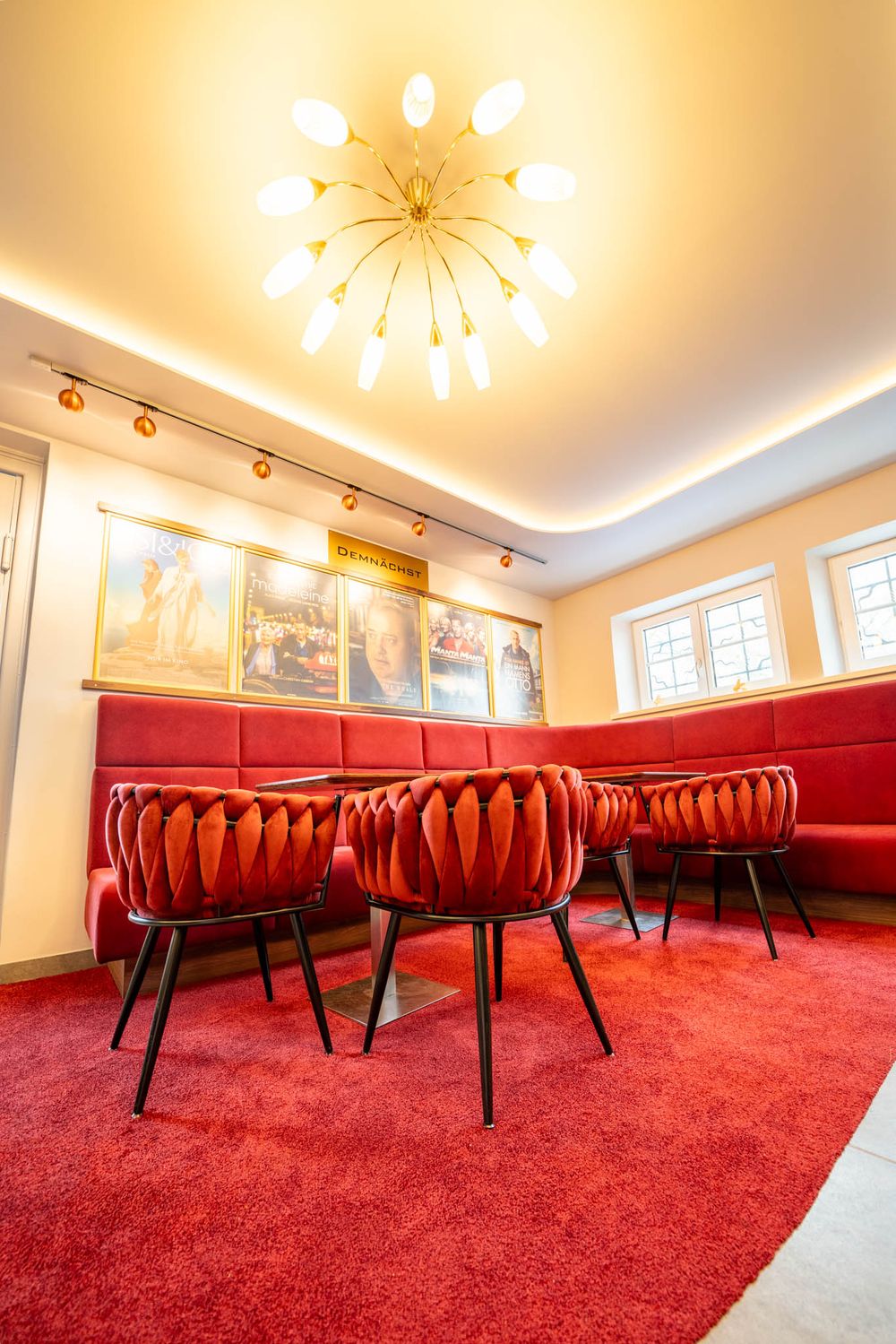 Modern restaurant interior with red velvet chairs and carpet, starburst chandelier, and framed artwork on cream walls.